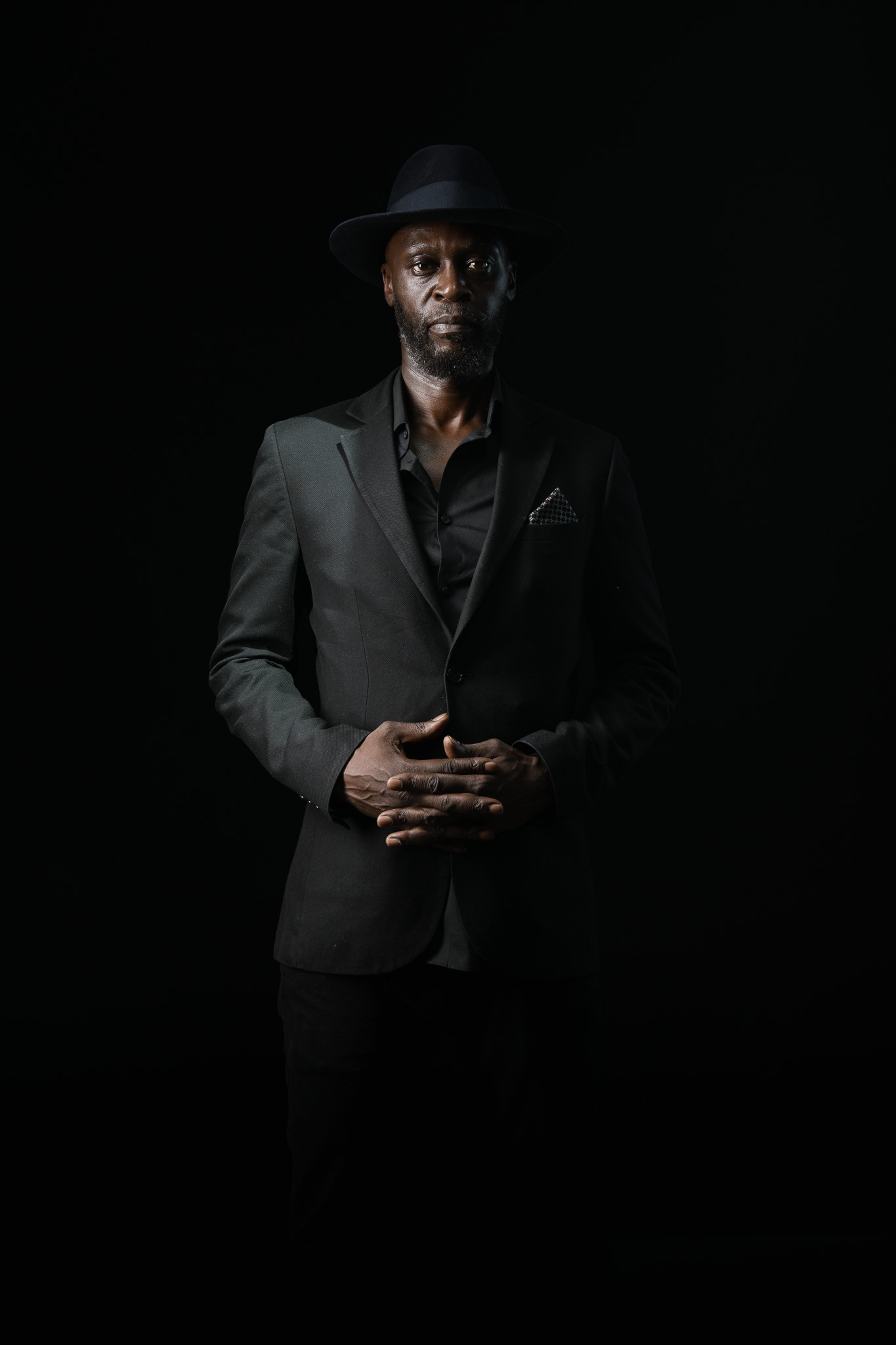 Portrait of Kyazi Lugangira in a black suit and hat against a dark background