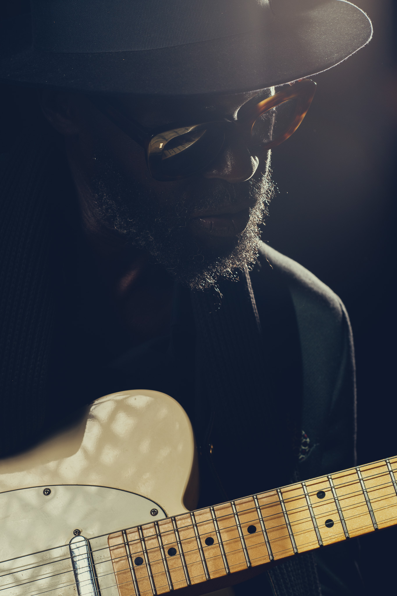 Close portrait of Kyazi Lugangira in hat and sunglasses holding an electric guitar in rim light