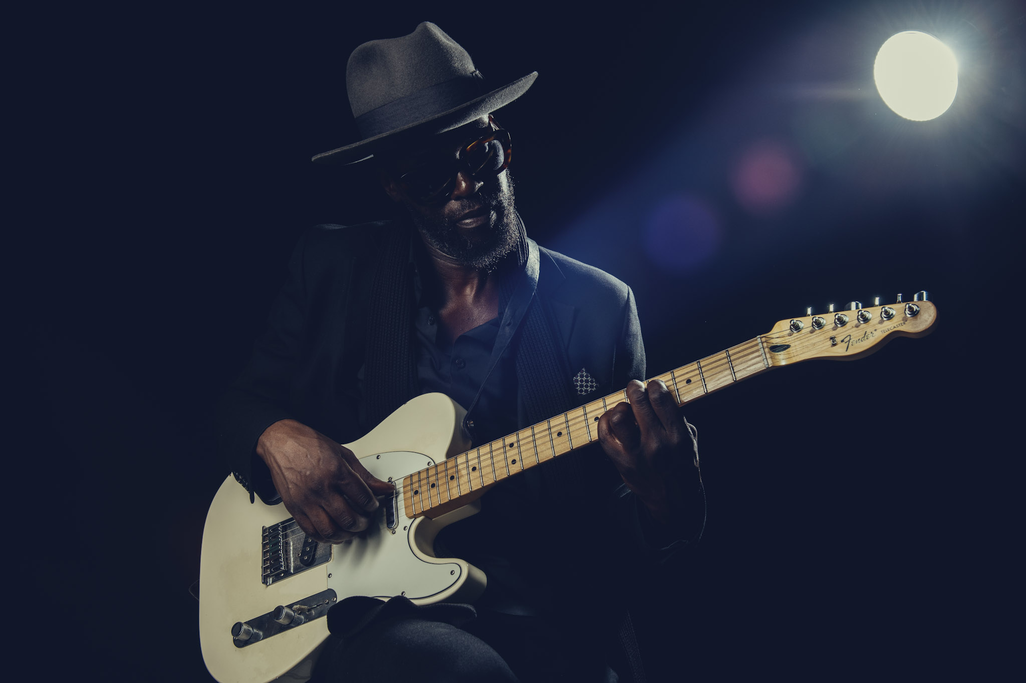 Kyazi Lugangira seated with a white Fender Telecaster beneath a circular studio light