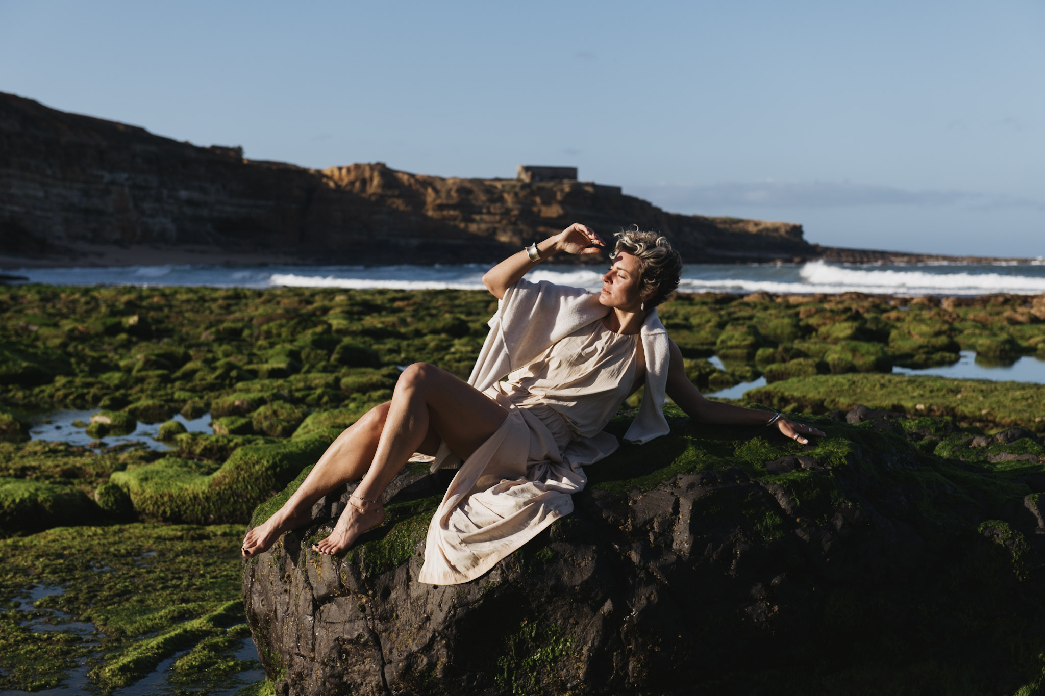 Woman in an ivory dress seated on moss-covered rocks beside the Atlantic surf in Portugal