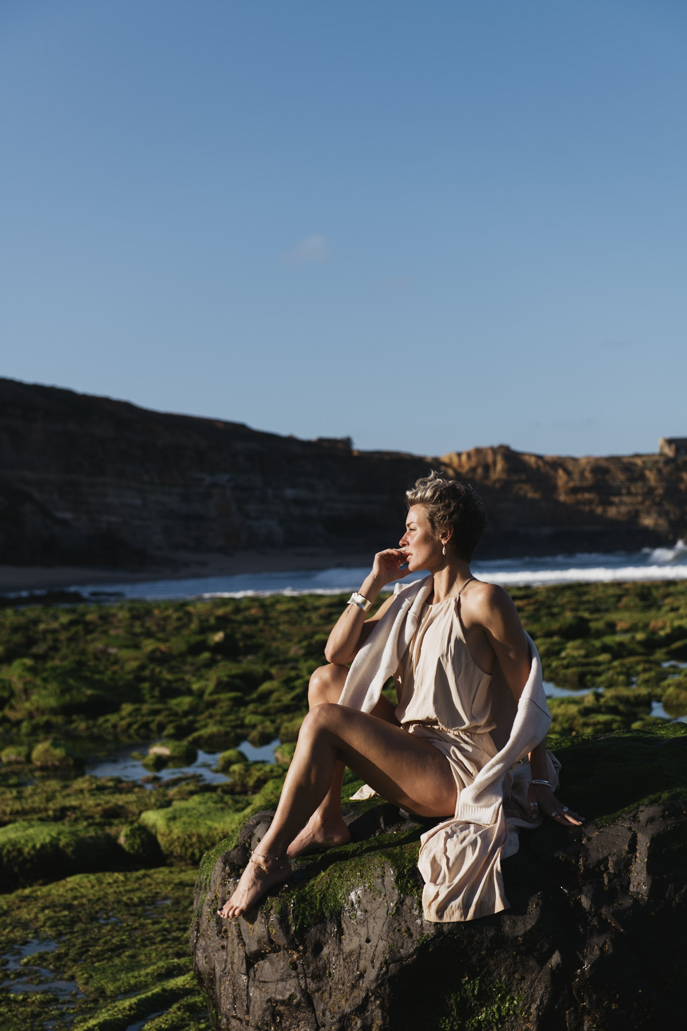 Vertical coastal portrait of a woman seated among tidal rocks beneath a clear blue sky