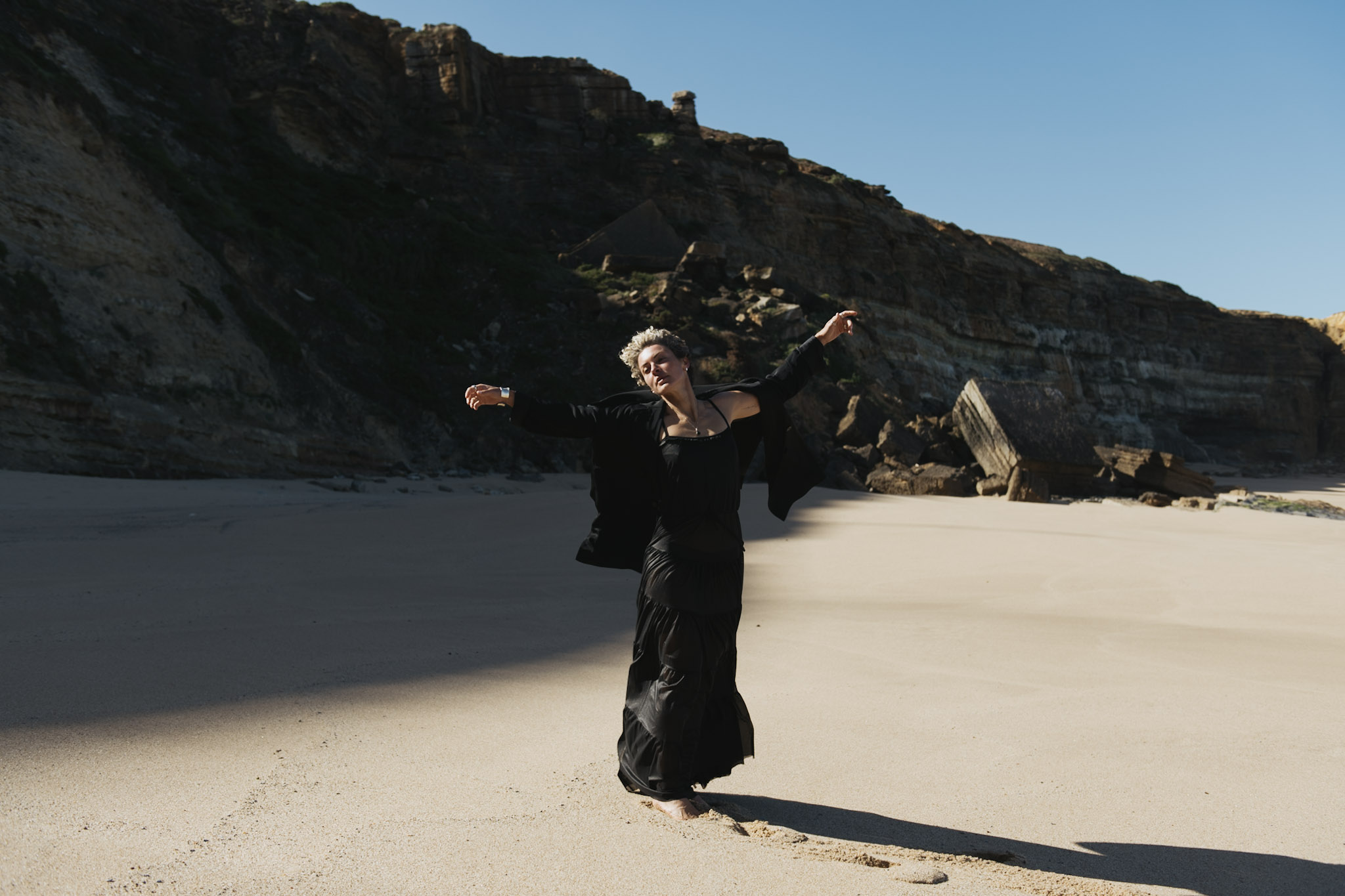 Woman in a flowing black dress moving across a cliff-lined beach
